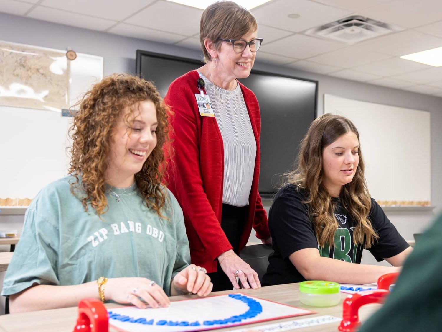 A professor in a red cardigan stands smiling behind two students seated at a classroom table. The student on the left laughs while arranging blue letter pieces on a board. She wears a “Zip Bang OBU” shirt, and the student on the right wears a black OBU shirt.