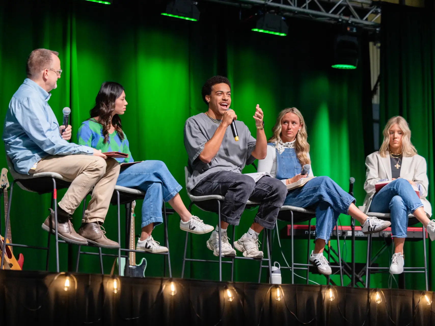 A panel of five people sitting on high stools on a stage with a vibrant green curtain backdrop. A young man in the center is speaking enthusiastically into a microphone, gesturing with his hands. The other panelists, including a man and three young women, listen intently. String lights decorate the front of the stage.
