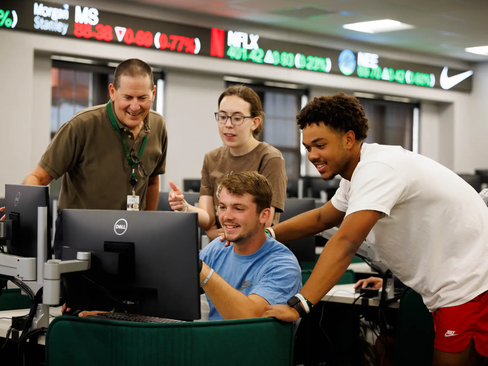 Four people gathered around a computer monitor in a modern finance lab. A young man sits at the desk while two other students and an older male instructor stand behind him, smiling and pointing at the screen. In the background, a digital stock ticker displays real-time data for companies like Netflix and Nike.