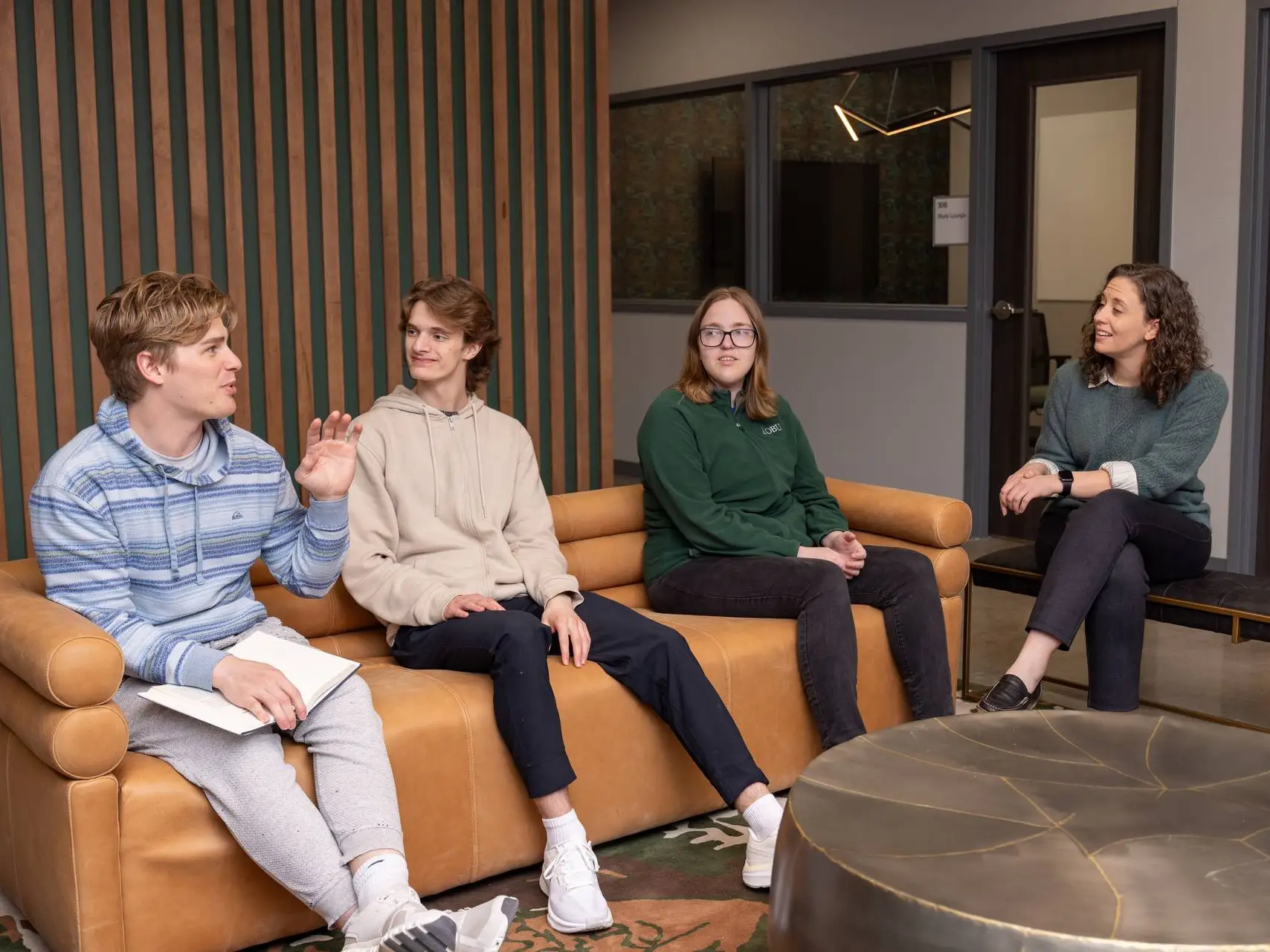 Four people engaged in a discussion while sitting in a modern lounge area. Three students sit on a tan leather sofa, while a woman sits on a bench opposite them. One student gestures while speaking, and the group maintains a relaxed, conversational atmosphere. The room features wood-slatted wall decor and a glass-walled office in the background.