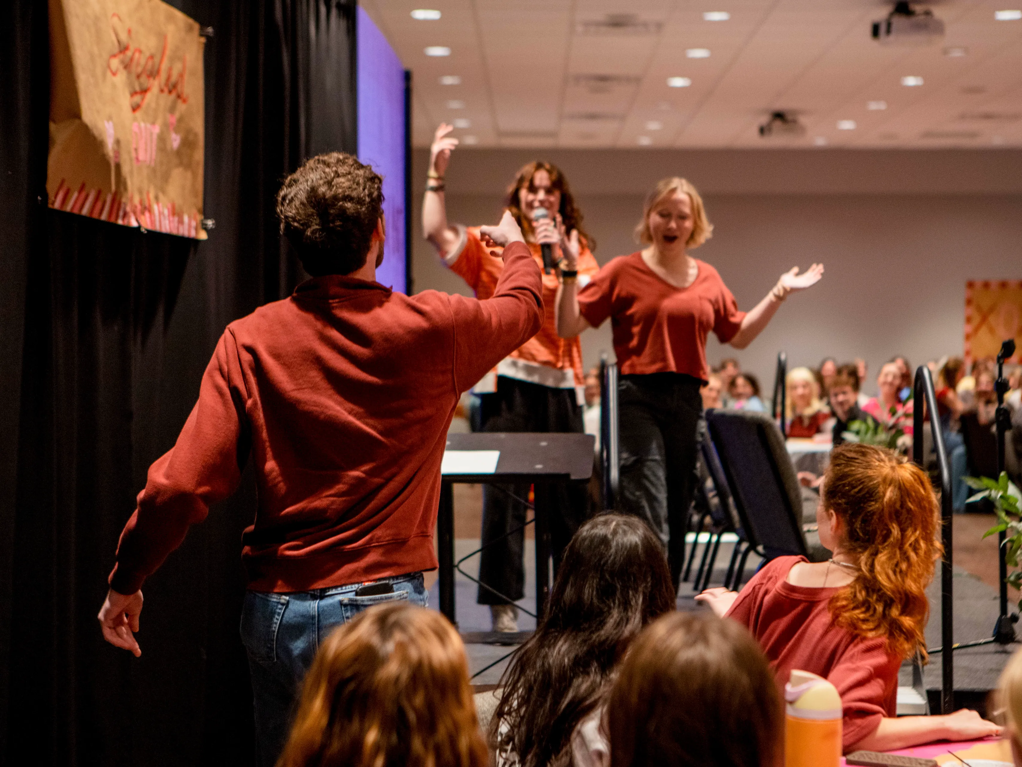 A student host in a rust-colored sweatshirt gestures toward two performers on stage in matching orange and red tops, one singing into a microphone with her arm raised, in front of a seated audience at round tables during an evening event.