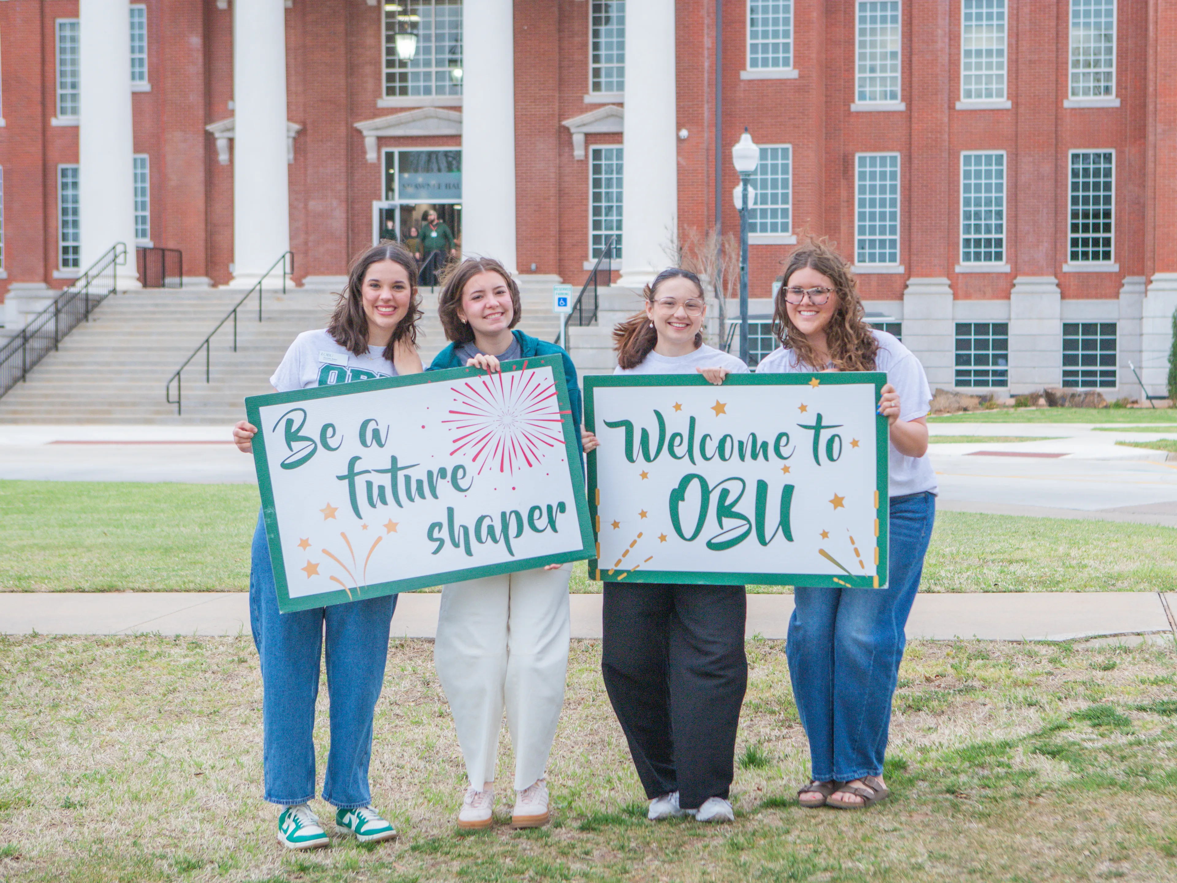 Four smiling students stand on a campus lawn holding signs that read "Be a future shaper" and "Welcome to OBU," with a brick building behind them.