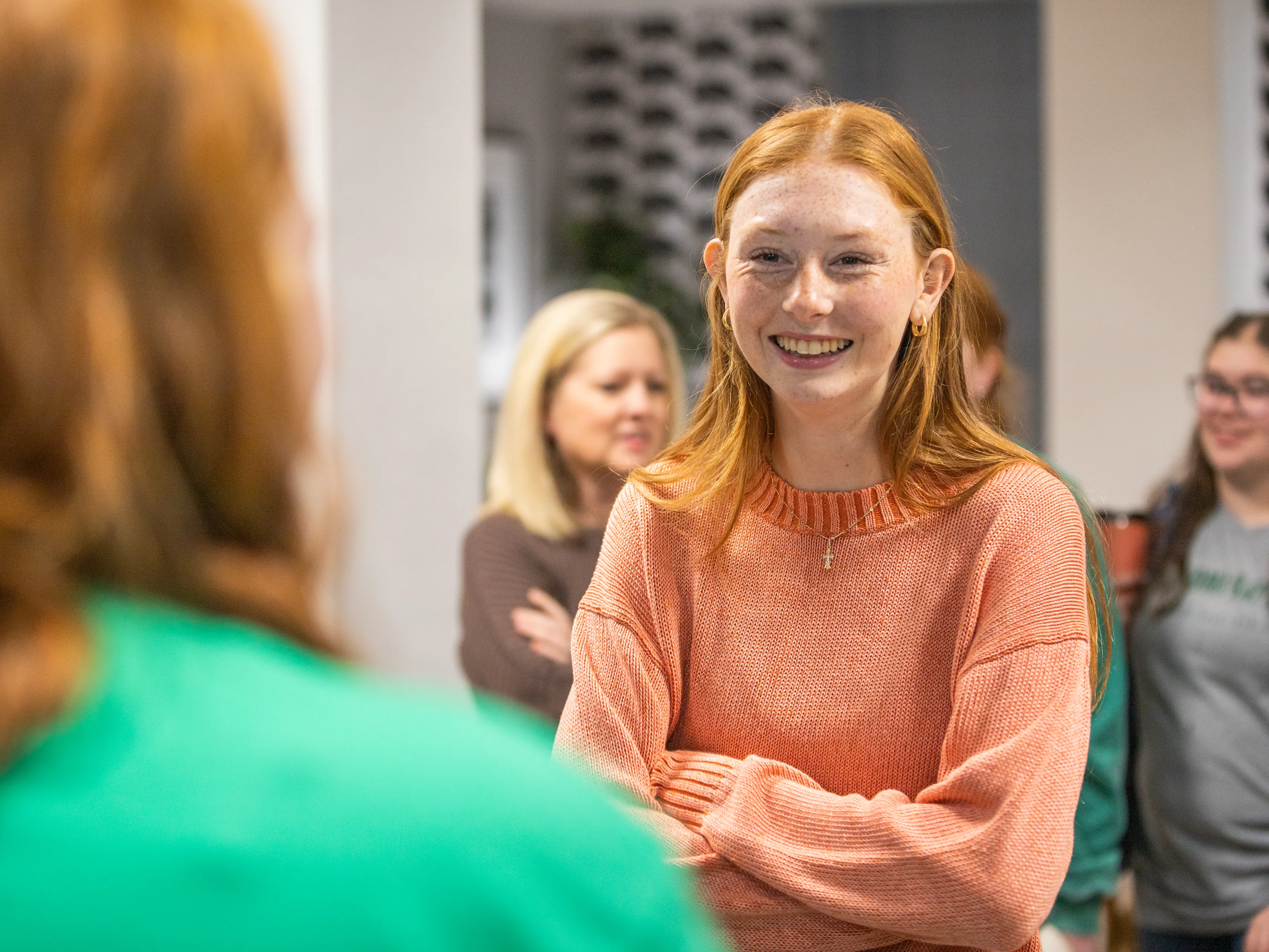 A smiling young woman with red hair and freckles, wearing an orange sweater and a small cross necklace, stands with her arms crossed during a campus visit, with other visitors blurred in the background.