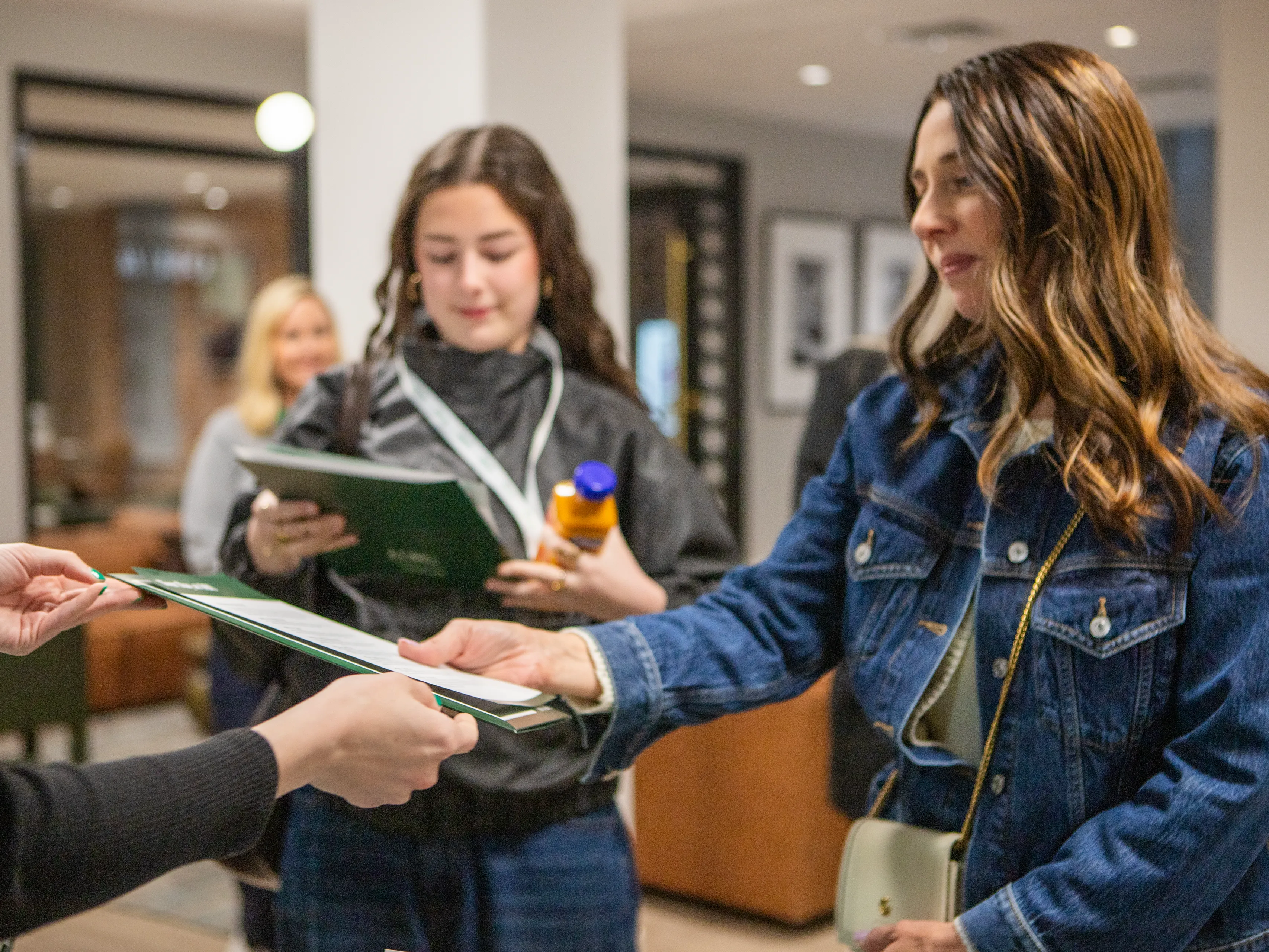 A staff member in a denim jacket hands a green folder of materials to another woman, while a student and another attendee look on nearby.