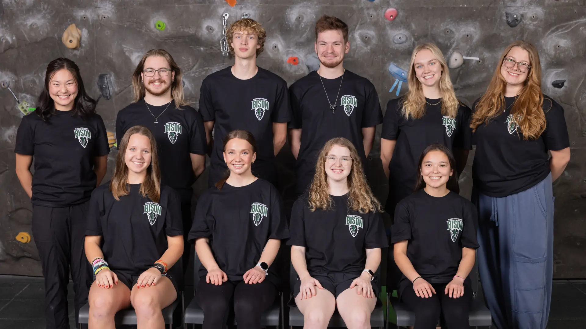 Ten members of the Climbing Club pose together in front of an indoor climbing wall, wearing black Bison Club Sports t-shirts.