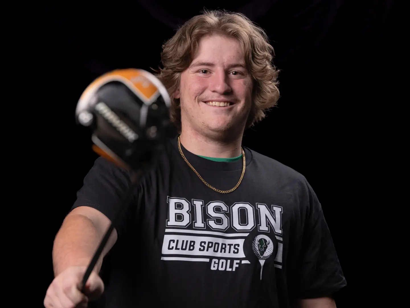 A smiling golfer in a black Bison Club Sports Golf t-shirt holds up a golf club, wearing a gold chain necklace.