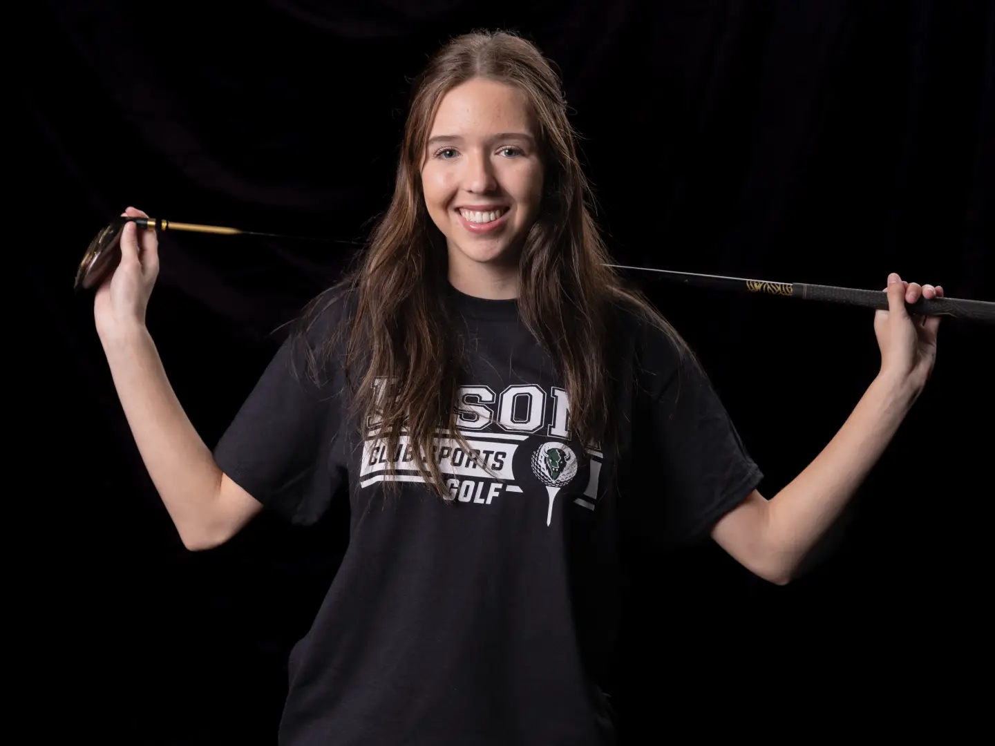 A smiling golfer holds two golf clubs up over her shoulders, wearing a black Bison Club Sports Golf t-shirt