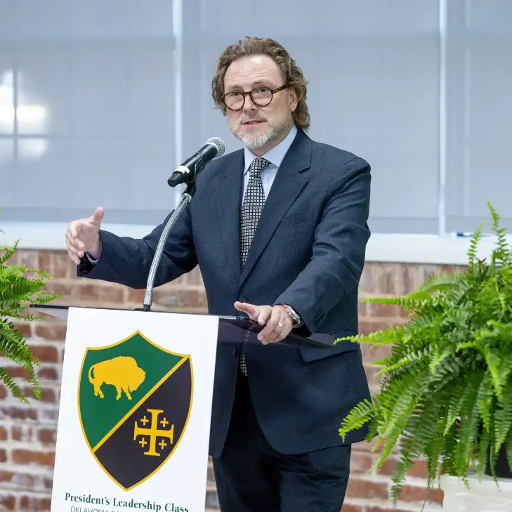 A man in glasses and a dark suit gestures while speaking at a podium bearing the OBU President's Leadership Class shield, flanked by potted ferns on pedestals in front of an exposed brick wall.