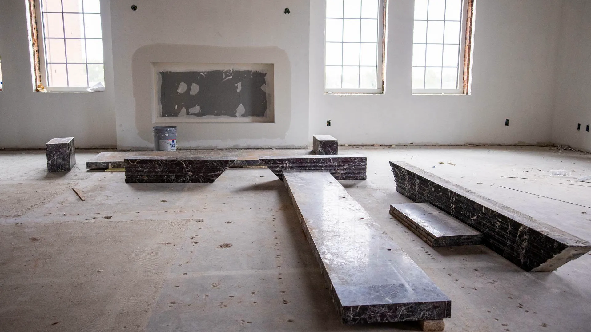 Interior room under construction with a drywalled fireplace niche flanked by windows, and dark marble slabs for the surround and hearth laid out unfinished on the concrete floor.
