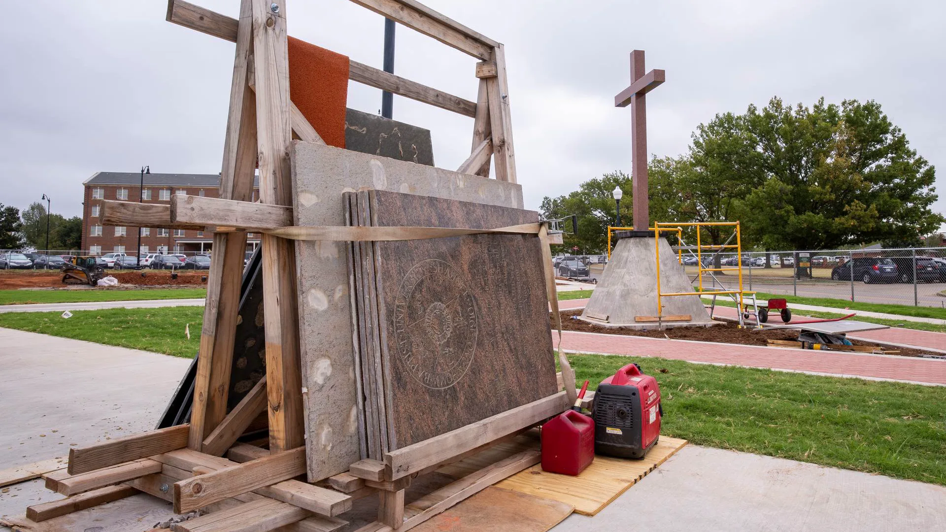 Engraved granite panel bearing the Oklahoma Baptist University seal, braced upright in a wooden frame outdoors, with a memorial cross monument and campus buildings visible in the background.