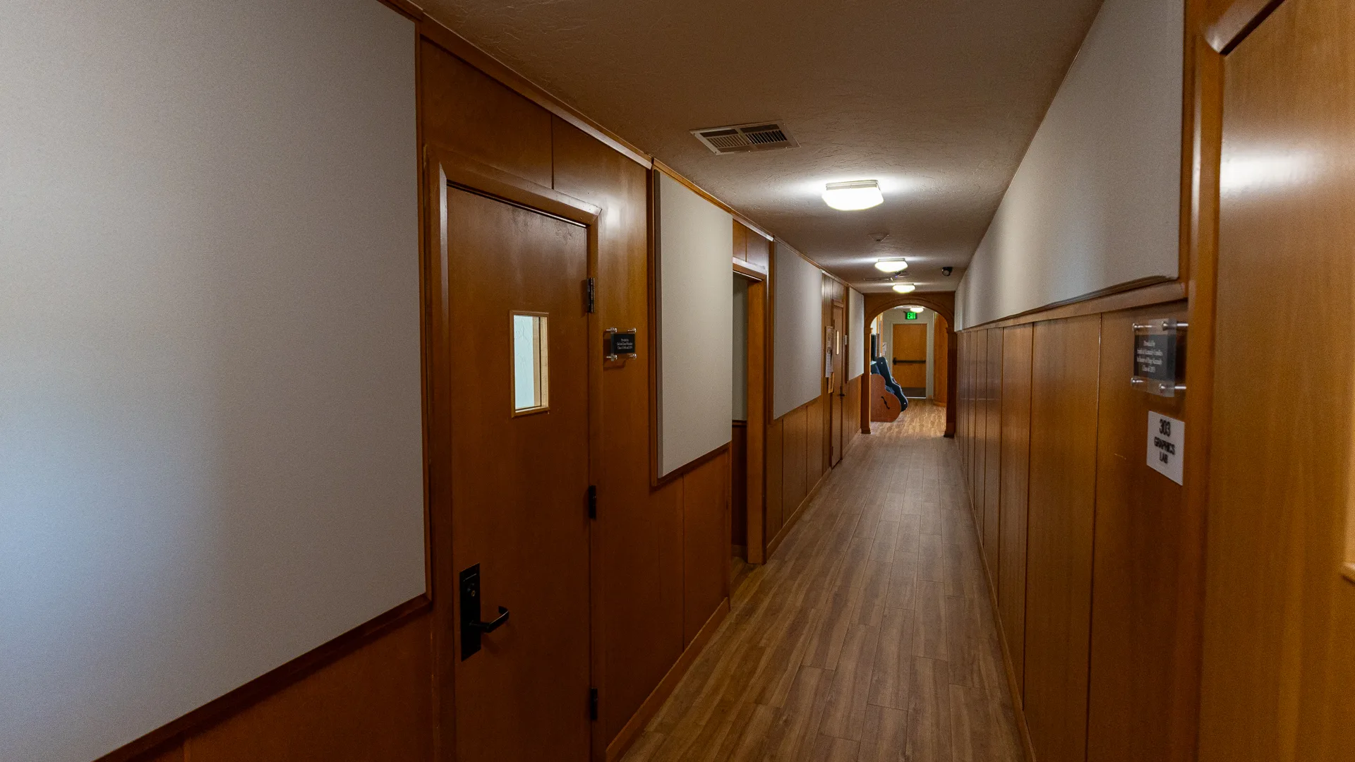 Wood-paneled hallway lined with office doors and nameplates, leading back to an archway and exit sign at the far end.