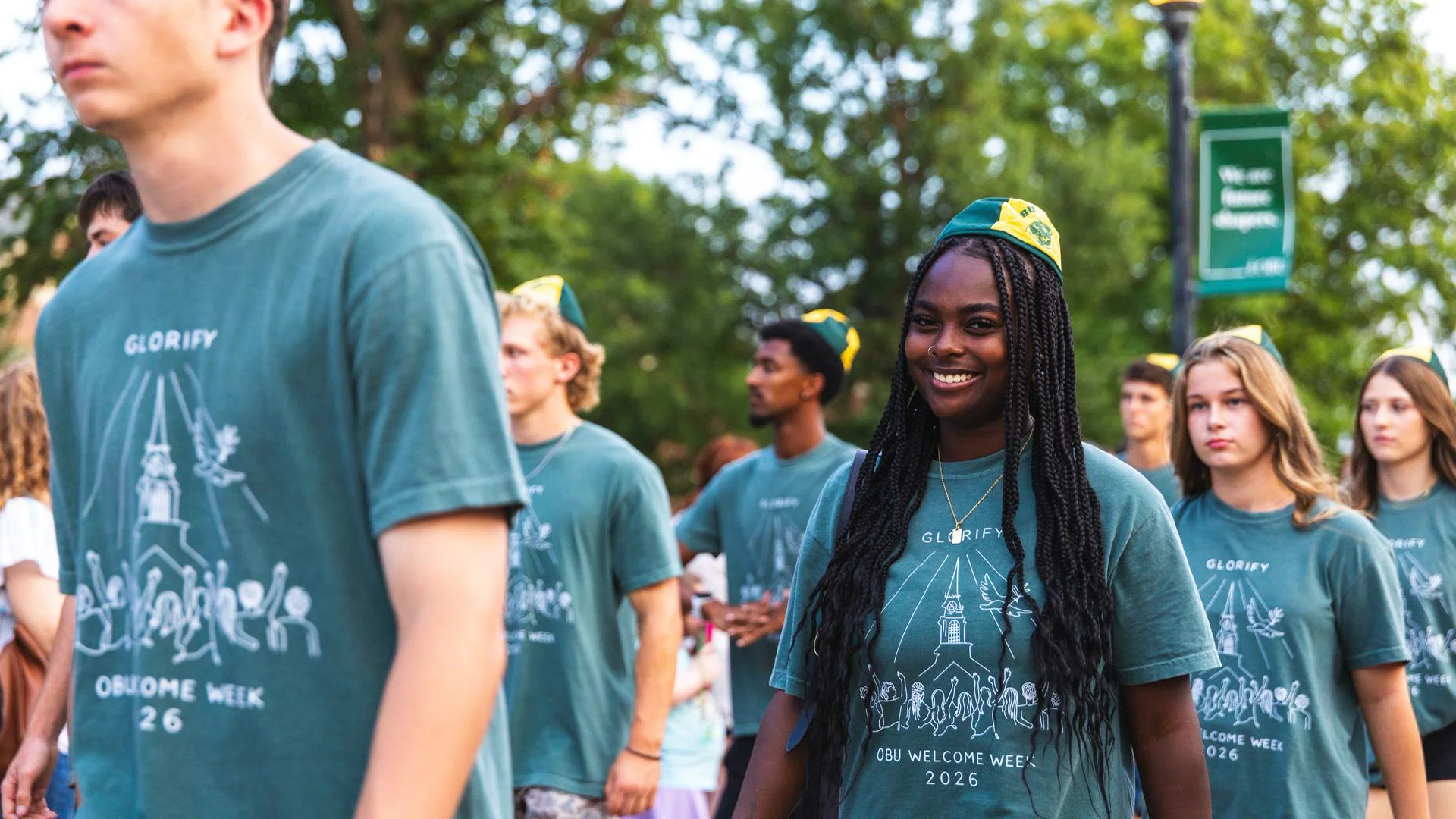 A smiling young woman with long braids and a green-and-yellow beanie walks with a crowd of students wearing matching "OBU Welcome Week 2026" T-shirts on a tree-lined campus path in the evening.