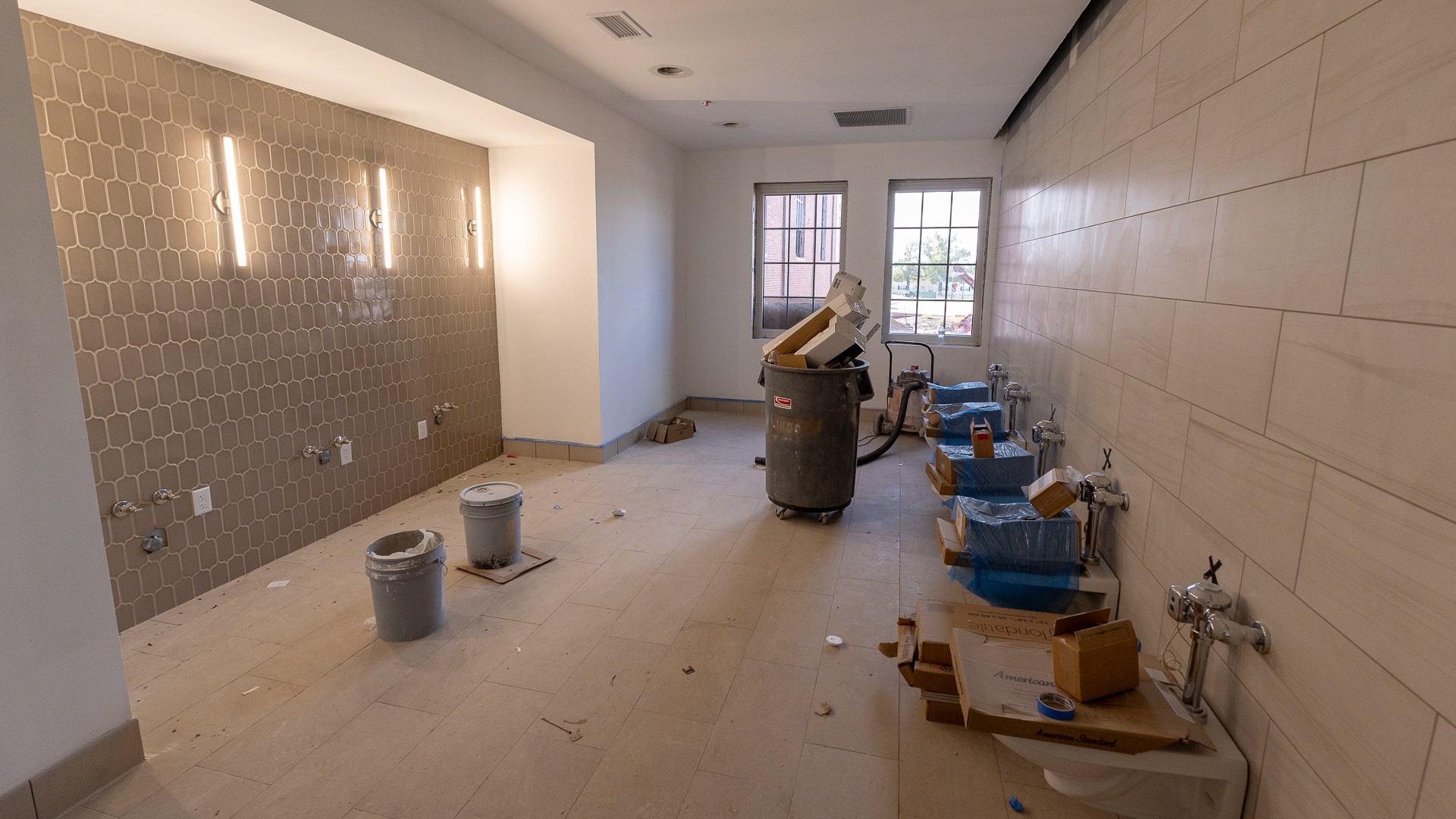 A restroom under construction, with a taupe tile accent wall and three lit vertical sconces on the left and wall-mounted toilets still wrapped in blue plastic along the tiled wall on the right. Buckets, boxes and a trash bin of debris sit on the new floor tile in front of two windows.