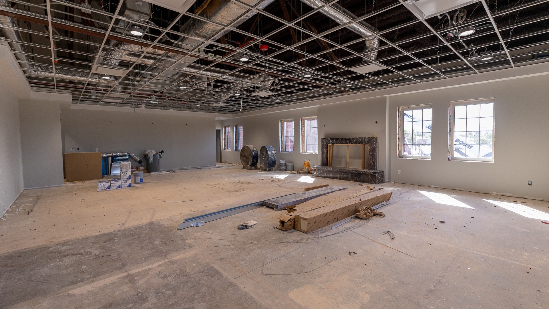 A large room under renovation with gray walls, an open ceiling grid exposing ductwork and lights, and a dark marble fireplace surround between rows of windows. Construction materials, fans and boxes are spread across the unfinished floor.