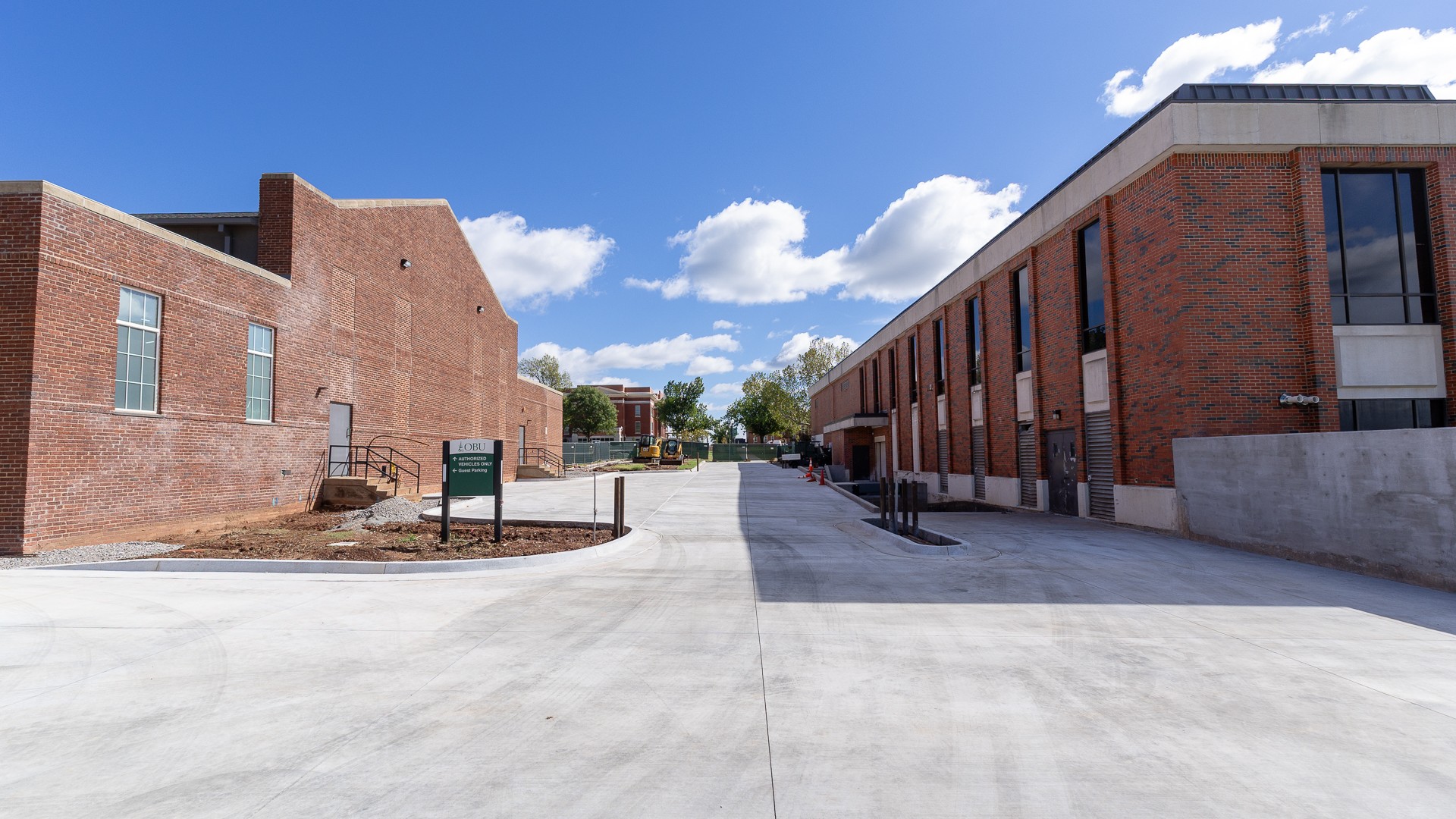 A newly poured concrete drive and loading dock between two red brick buildings under a blue sky with scattered clouds. A green OBU sign directs authorized vehicles straight ahead and guest parking to the left, with construction equipment and fencing in the distance.