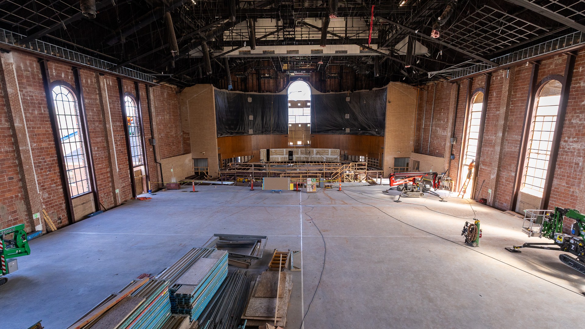 A wide view from above of a cavernous hall under renovation, with tall arched windows along both brick side walls and an arched window centered above a stage area at the far end. Lifts, stacked materials and cords sit on the bare concrete floor slab.