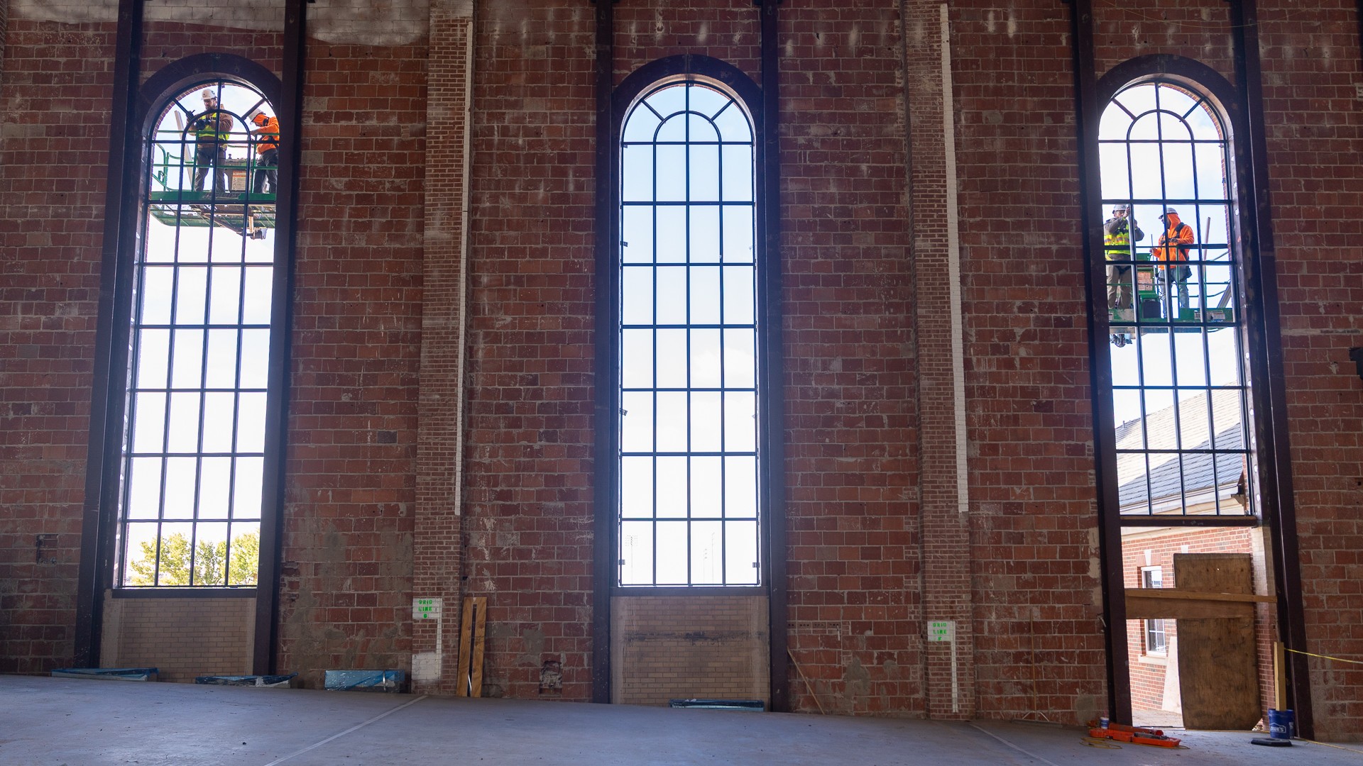 Three tall arched window frames in an exposed brick wall, viewed from inside the building. Through the empty frames, construction workers in hard hats and safety vests stand on lifts outside.