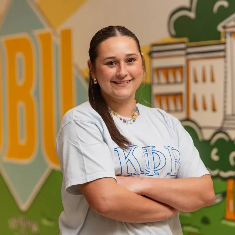 Portrait photo of Chesney Long. Female student in a gray and green tee-shirt standing in front of a mural wall.