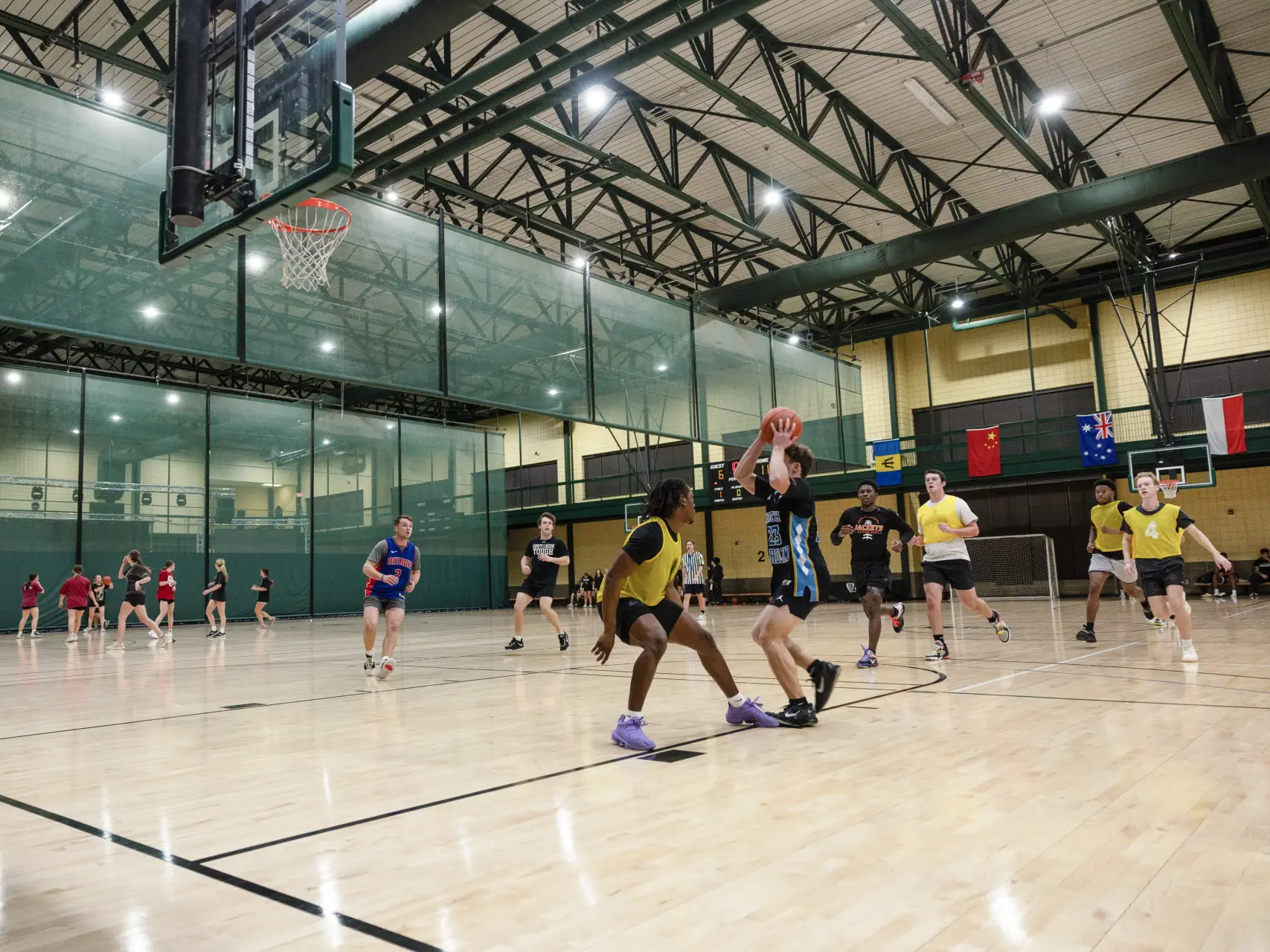 An action shot of an indoor intramural basketball game on a hardwood court. A player in a black and blue jersey prepares to shoot the ball while being guarded by a player in a yellow pinnie. Other students in various athletic gear are spread across the court, and a separate group is visible playing behind a green mesh divider in the background. Several international flags hang from the far balcony of the gymnasium.