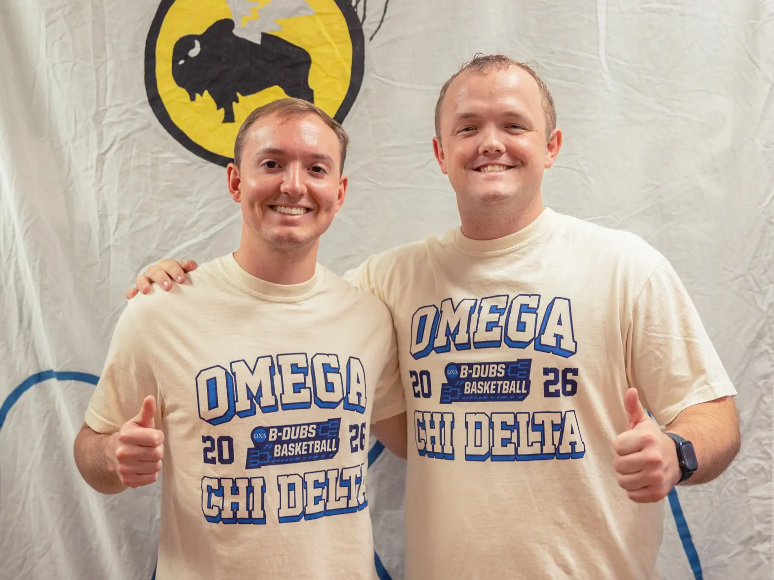 Two young men standing side-by-side, giving a thumbs-up to the camera. They are wearing cream-colored t-shirts that say "OMEGA CHI DELTA" in blue block letters. Between the words, there is a small logo that reads "B-Dubs Basketball 2026." Behind them is a white banner featuring a yellow circular logo of a buffalo with wings.