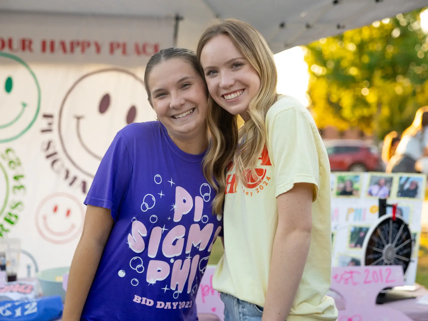 Two young women smiling closely together under a white tent. The woman on the left wears a purple shirt that says "PI SIGMA PHI BID DAY 2023" in pink letters. The woman on the right wears a pale yellow shirt. In the background, there is a banner with smiley faces and a "Pi Sigma Phi" display board.