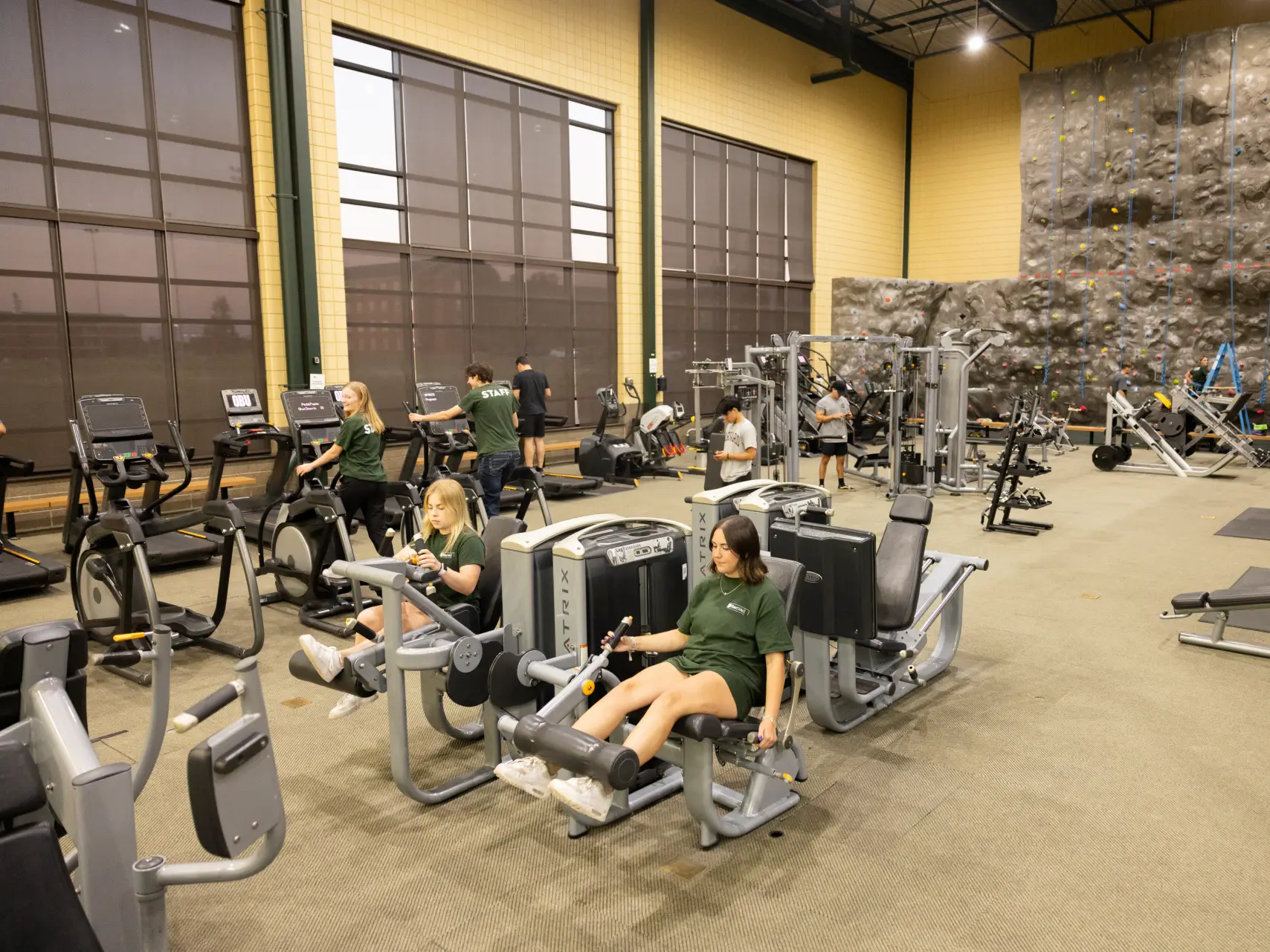The interior of a spacious, high-ceilinged campus gym. Several students are using various exercise machines, including treadmills and leg presses. In the background, a large, multi-story indoor rock climbing wall spans the far wall.