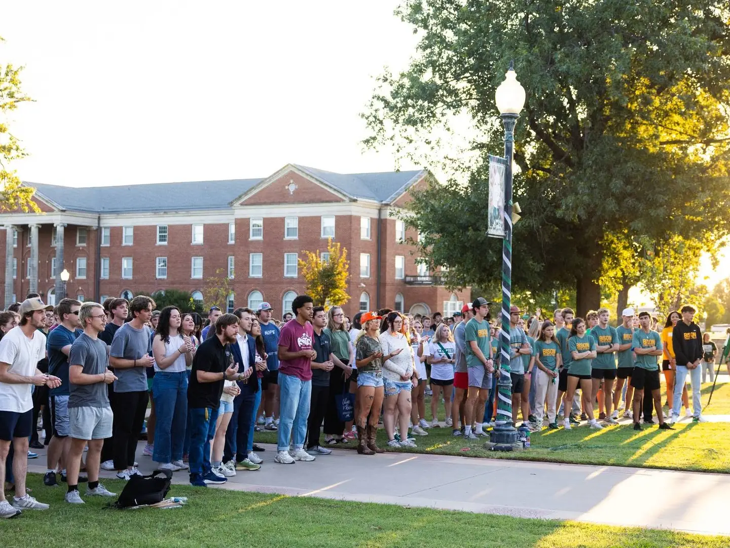 A large crowd of students gathered on a grassy campus lawn during golden hour. They are standing attentively, looking toward an off-camera speaker. A brick campus building with white columns is visible in the background.