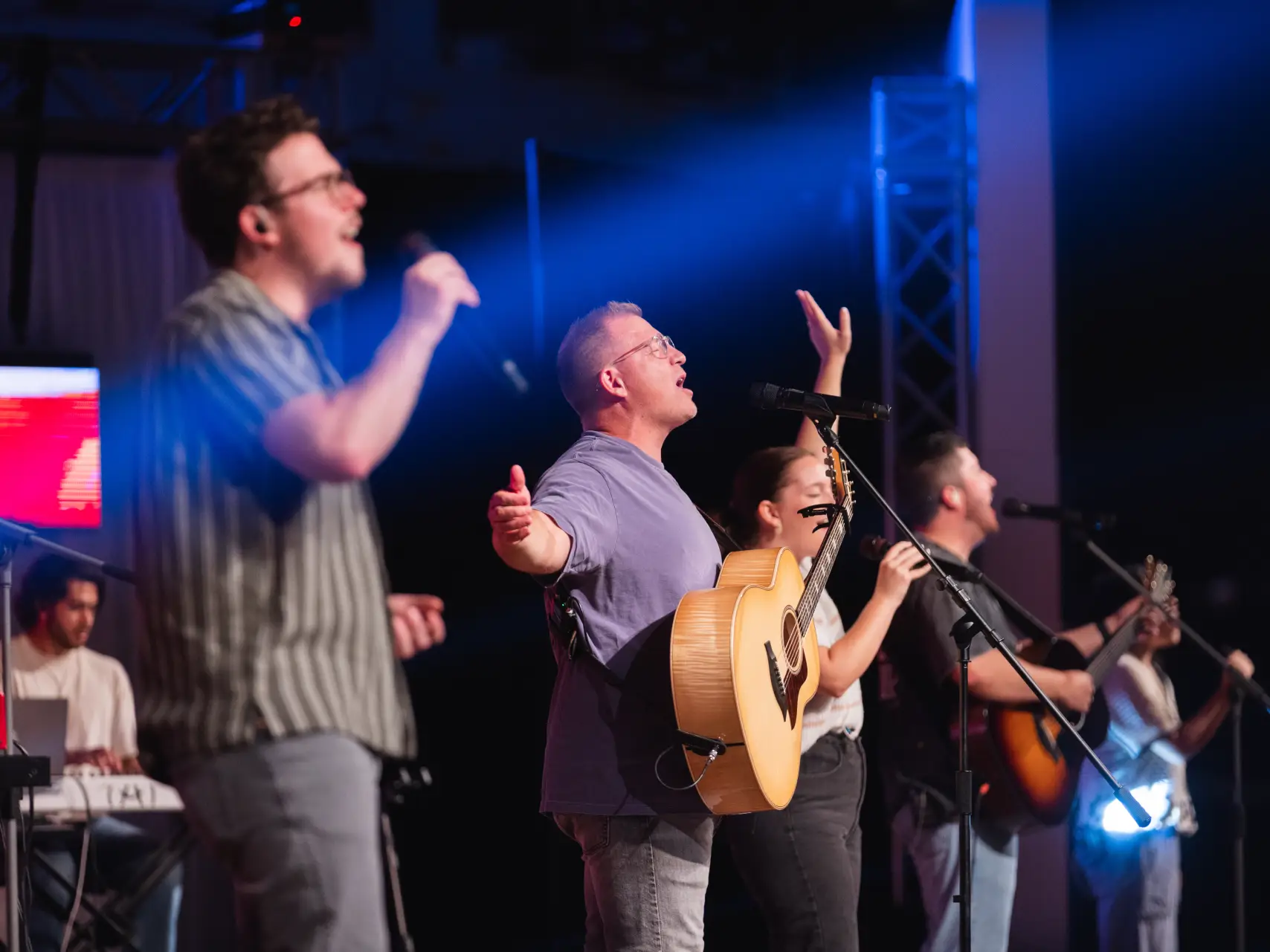 A worship band performing on a stage under blue spotlights. Three vocalists are singing passionately into microphones; the central singer is playing an acoustic guitar. A keyboardist is partially visible in the background.