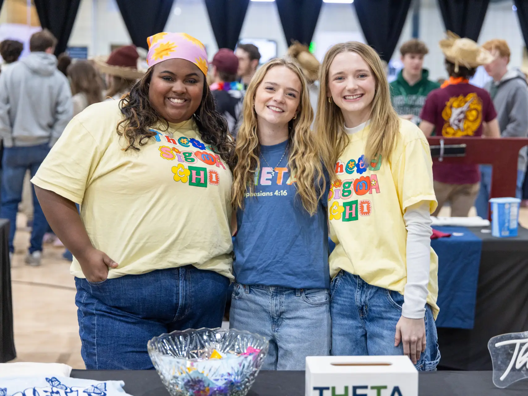 Three young women smiling behind a table at an indoor event. The two women on the ends are wearing pale yellow t-shirts with "Theta Sigma Chi" printed in colorful, flowery bubble letters. The woman in the center is wearing a blue t-shirt with "THETA" printed on it. A clear bowl of candy and a sign with "THETA" are on the table in the foreground.