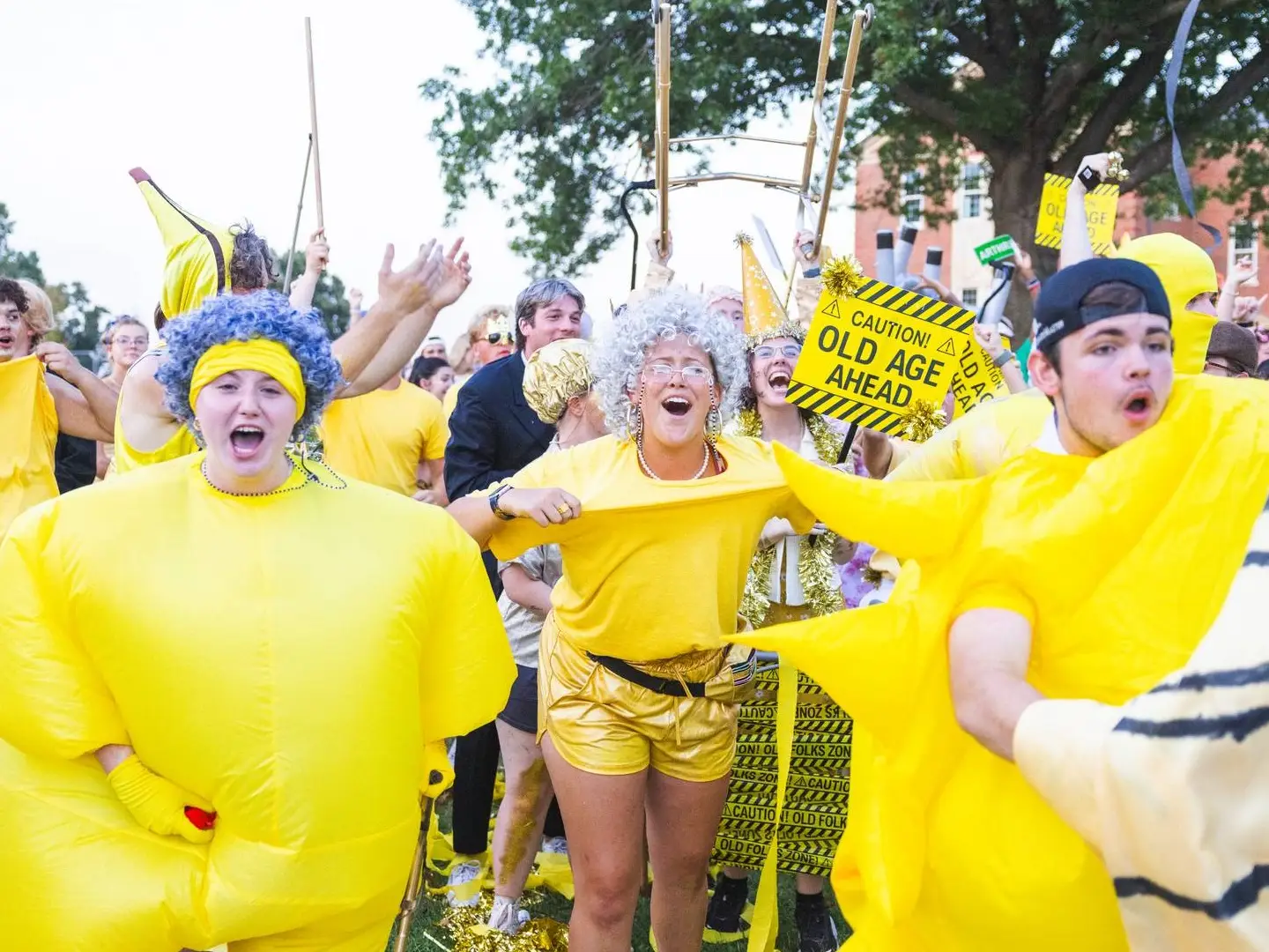A group of students dressed in bright yellow costumes and props, celebrating a campus tradition. One student wears a large inflatable yellow suit, another wears a silver wig and glasses with a "Caution! Old Age Ahead" sign, and others are cheering in the background.