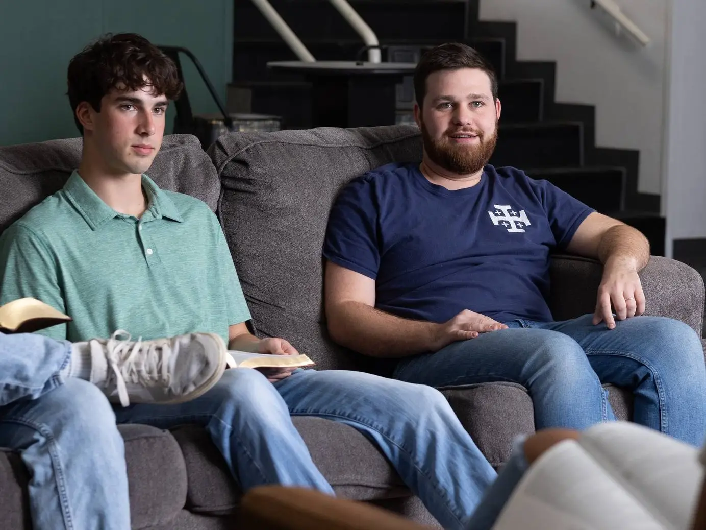 Two young men are seated on a grey sofa during a group meeting. The man on the left wears a light green polo shirt and holds an open Bible, while the man on the right wears a navy blue T-shirt with a white cross emblem and is looking toward someone off-camera.