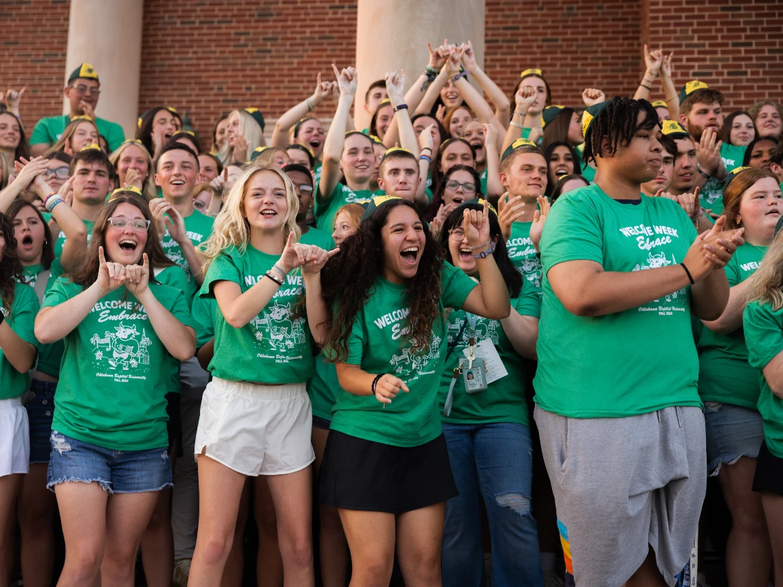 Two smiling students, a man in a grey "Oklahoma Baptist University" hoodie and a woman in a black puffer jacket, stand outdoors in front of a brick campus building. They are both making a "Bison" hand sign with their pinky and index fingers extended.