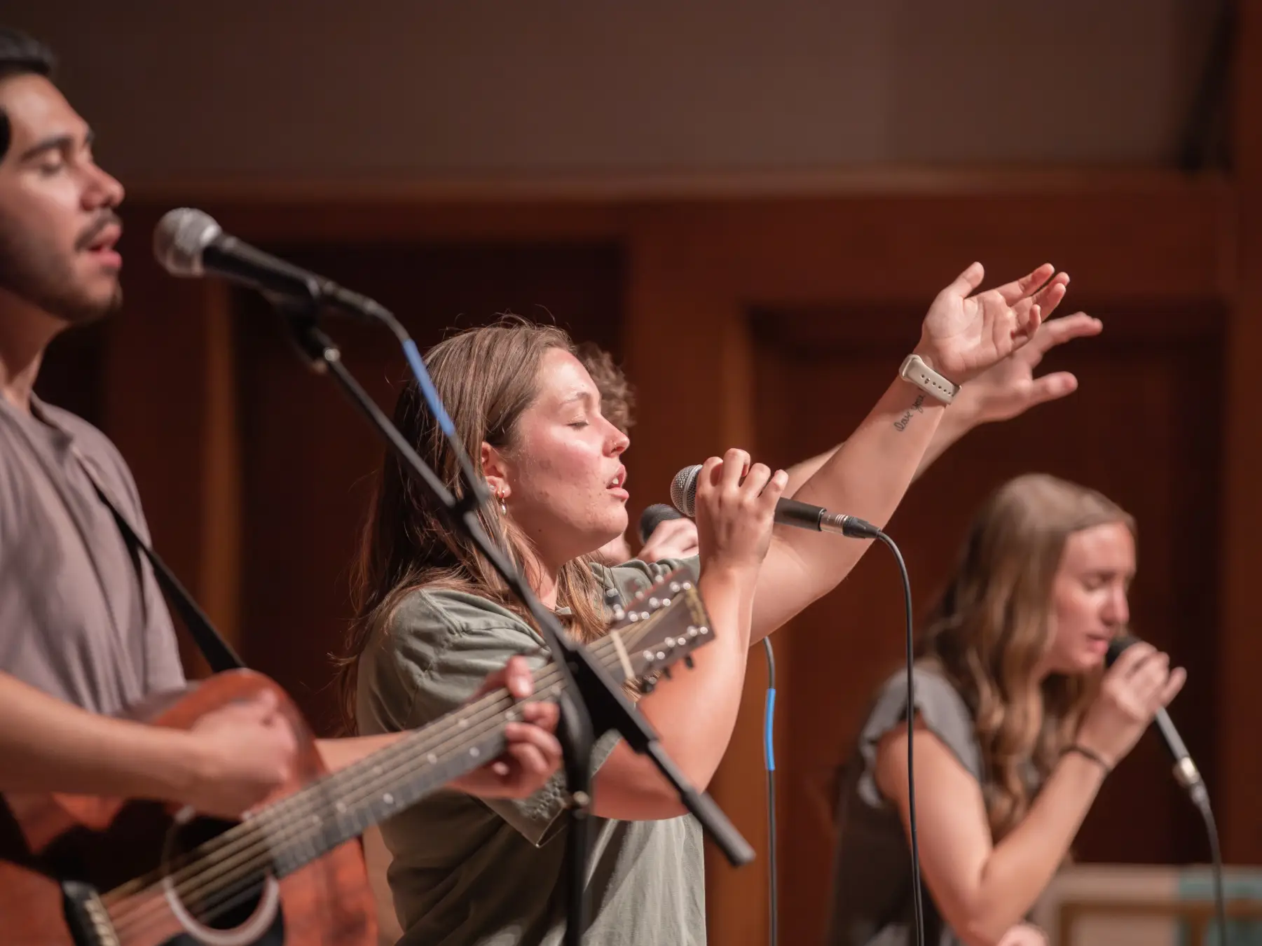 A close-up of a worship band performing. A woman in the center sings passionately into a microphone with one hand raised. To her left, a man plays an acoustic guitar, and another woman sings in the background.