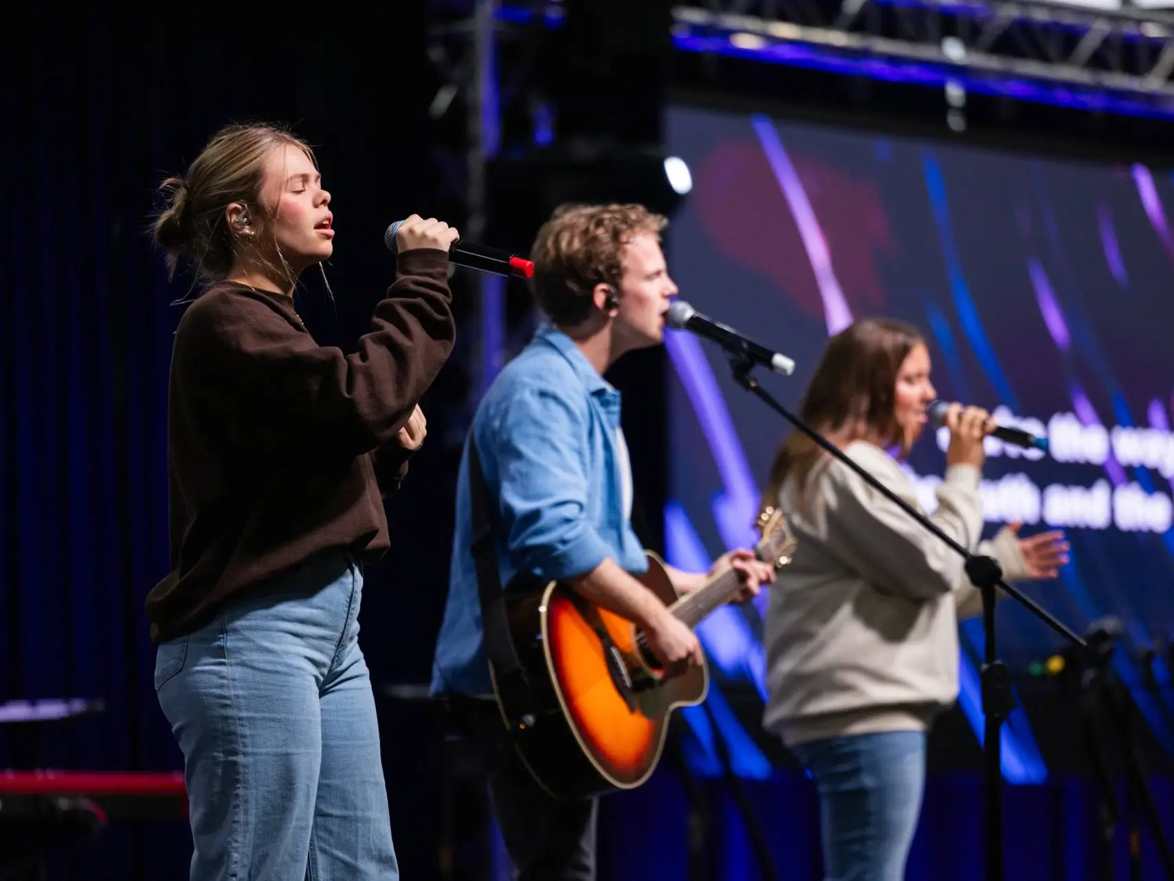 An image shows three people performing on a stage. In the foreground, a woman with hair in a bun wears a brown sweatshirt and light blue jeans, singing into a handheld microphone with her eyes closed. In the center, a man in a blue button-down shirt plays an acoustic guitar and sings into a microphone on a stand. In the background, another woman in a light-colored sweatshirt sings into a microphone. They are on a stage with a dark background and a large screen displaying blue and purple abstract graphics with some lyrics visible.
