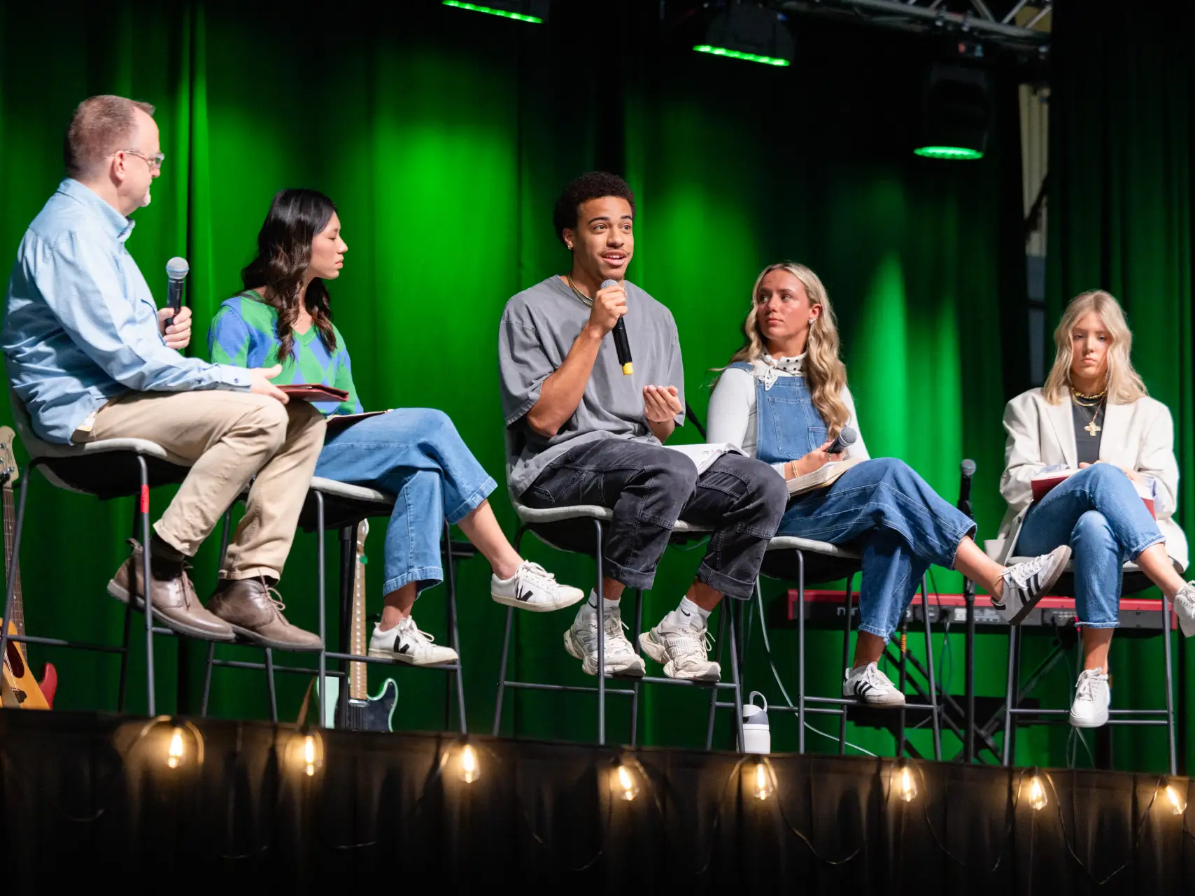 An image shows five people sitting on a stage in front of a green curtain. From left to right, a man in a light blue button-up shirt and tan pants sits holding a microphone. Next to him, a woman in a green and blue patterned sweater and jeans looks toward the center. In the middle, a man in a grey t-shirt and dark pants speaks into a microphone. To his right, a woman in a denim jumper and white shirt sits holding a microphone. On the far right, a woman in a light grey blazer and jeans sits looking down at a notebook. String lights are visible along the bottom of the frame.