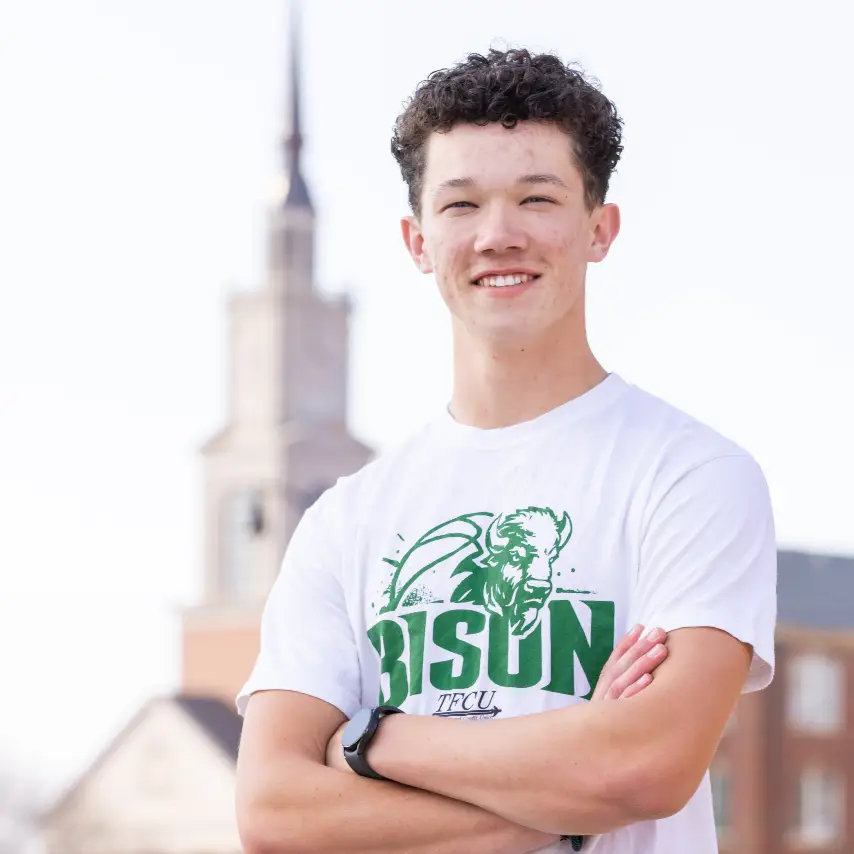 A portrait of a smiling young man with curly hair, Judah Motley, wearing a white T-shirt with a green "Bison" logo. He has his arms crossed, and a blurred white chapel tower is visible in the background under a bright sky.