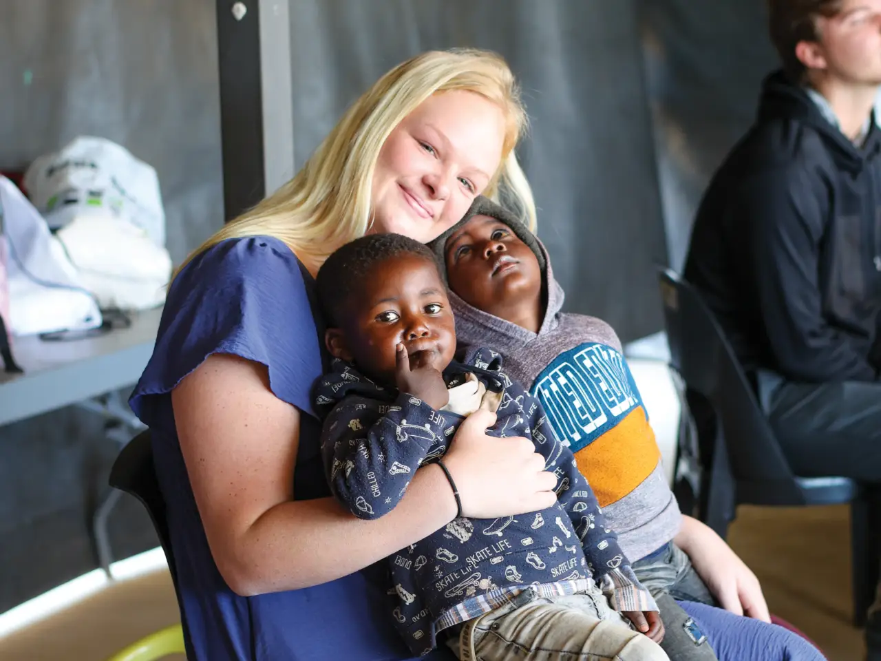 A smiling young woman in a blue top sits and holds two young children in her lap. One child looks directly at the camera with a finger to their lips, while the other leans back against her, looking upward.