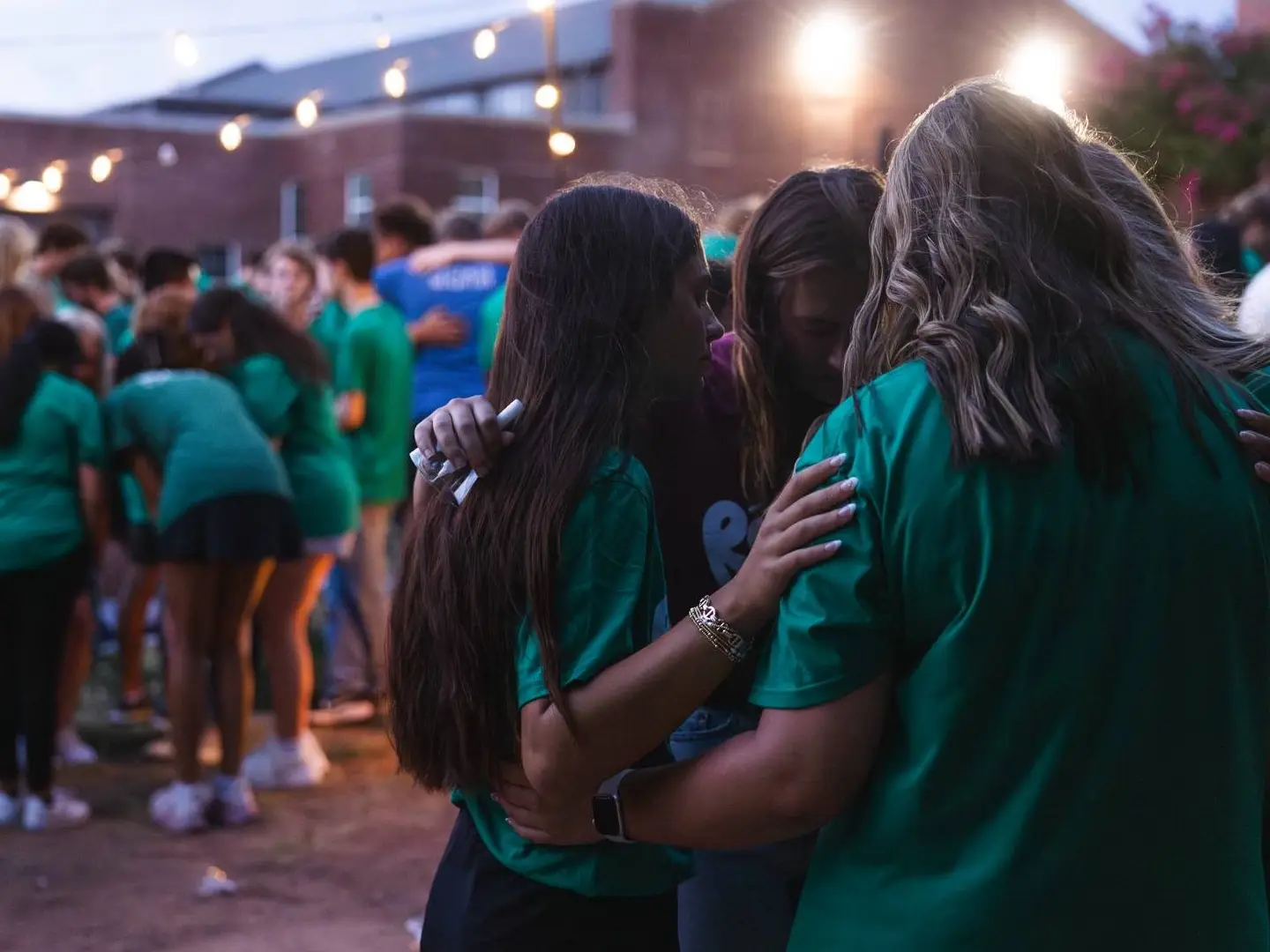 A group of students in matching green shirts gather outdoors at dusk under hanging string lights. In the foreground, three students are huddled closely together with their hands on each other's shoulders in a moment of prayer or reflection.