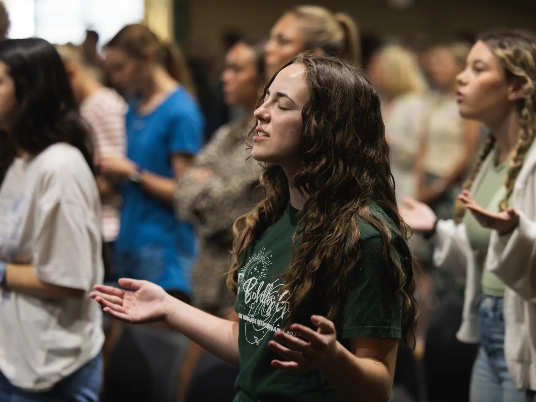 A young woman with long, wavy hair stands with her eyes closed and hands held out in a gesture of worship. She is wearing a dark green graphic t-shirt and is surrounded by a blurred crowd of people in a similar setting.
